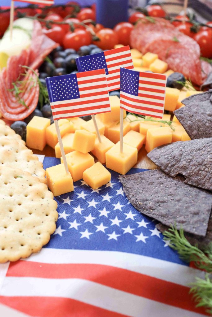A patriotic 4th of July Charcuterie Board featuring American flag toothpicks, cheddar cubes, crackers, blue corn chips, tomatoes, salami, and blueberries arranged on an American flag-themed cloth.