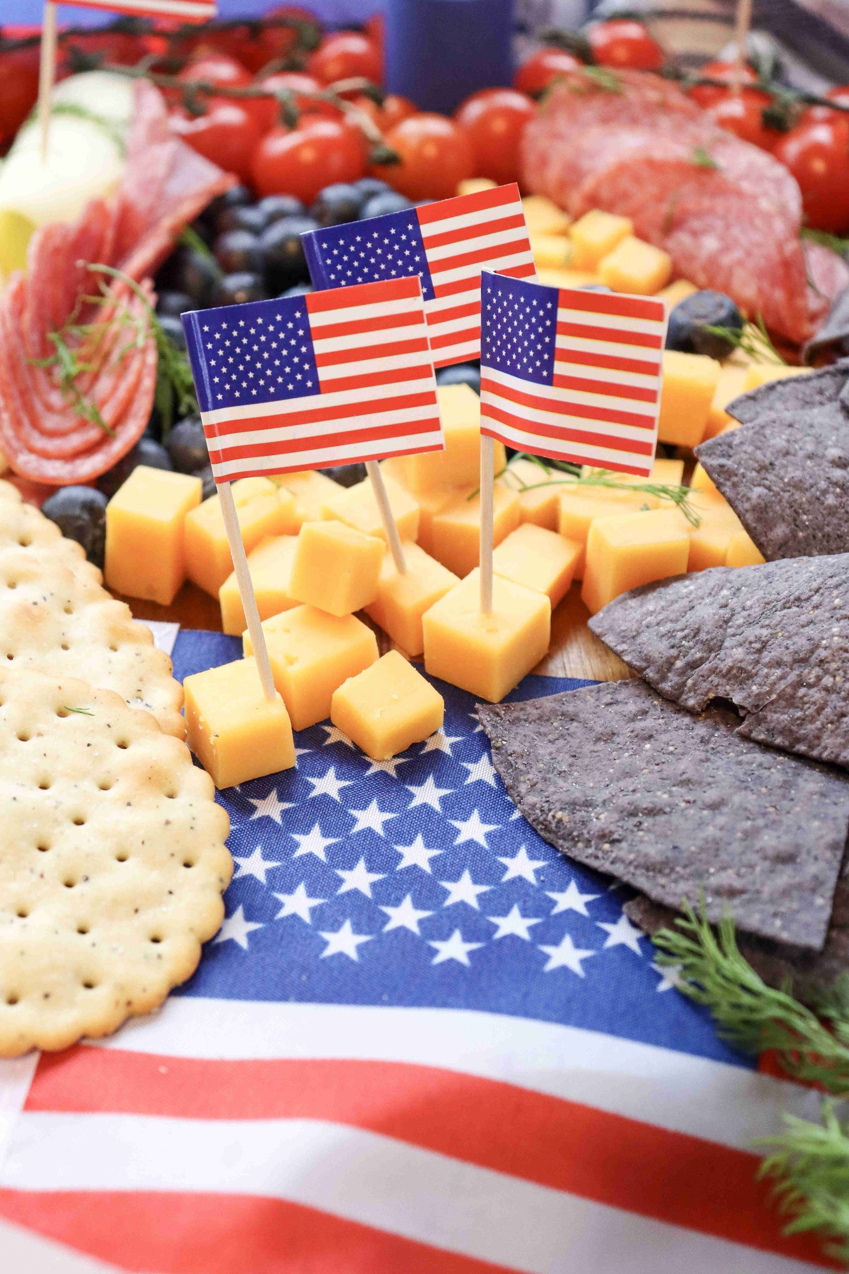 A patriotic 4th of July Charcuterie Board featuring American flag toothpicks, cheddar cubes, crackers, blue corn chips, tomatoes, salami, and blueberries arranged on an American flag-themed cloth.