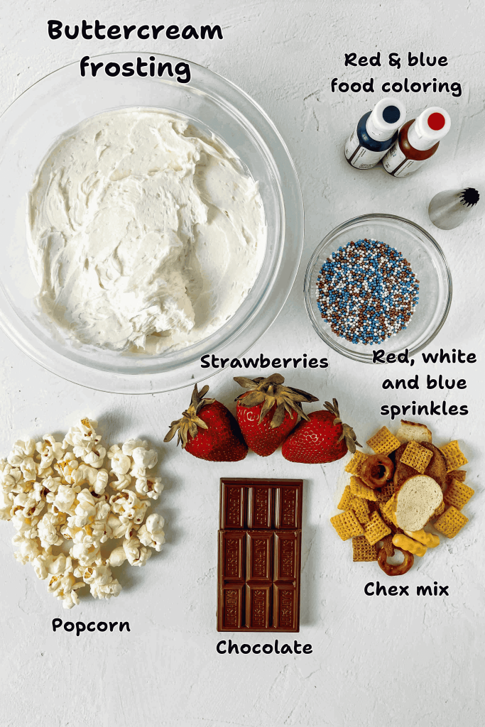 A festive flat lay perfect for a 4th of July Charcuterie Board: buttercream frosting, red and blue food coloring, patriotic sprinkles, strawberries, popcorn, a chocolate bar, and Chex mix on a white surface.