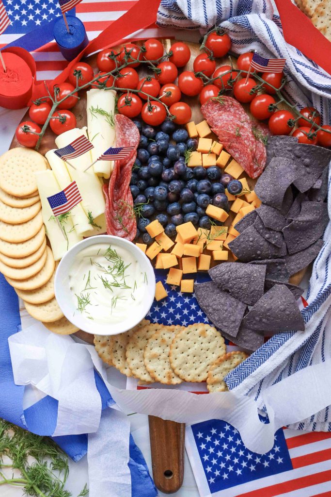 A festive 4th of July Charcuterie Board featuring red tomatoes, blueberries, cheese cubes, crackers, blue corn chips, salami, string cheese with small American flags, dip, and napkins—all decorated with patriotic flag motifs.