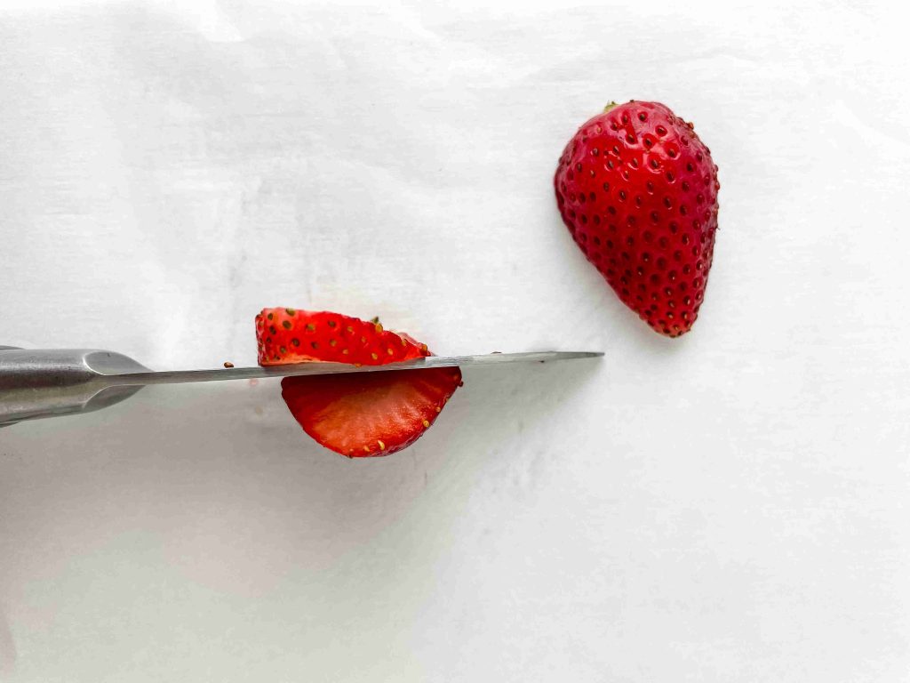 A knife slices through a fresh strawberry on a white surface, perfect for adding to your 4th of July Charcuterie Board—with one half already cut and the other still intact.