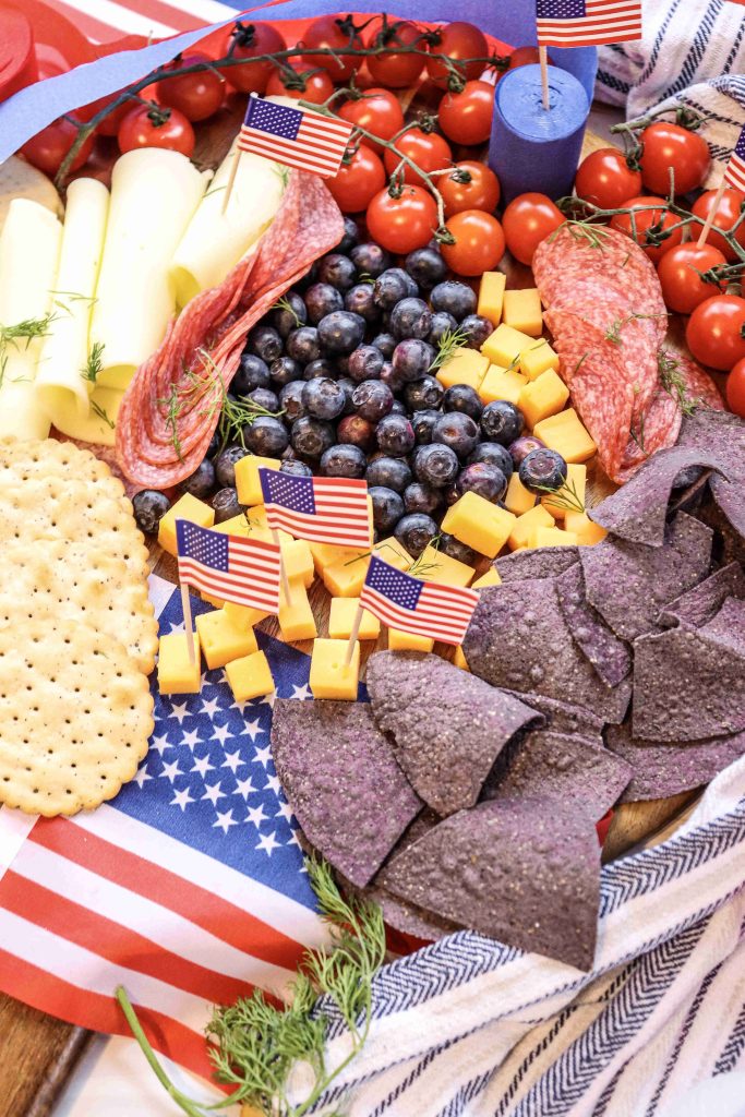 A festive 4th of July Charcuterie Board features yellow cheese cubes, blueberries, grape tomatoes, rolled cheese, salami, crackers, blue corn chips, and mini American flags, all beautifully arranged on a patriotic napkin.