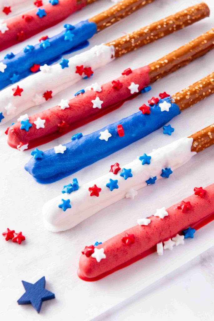 Red White and Blue Pretzel Rods dipped in candy melts and topped with star-shaped sprinkles make perfect patriotic pretzel rods, arranged on a white surface for festive 4th of July snacks.