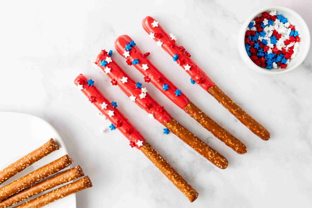 Four patriotic pretzel rods dipped in red coating and decorated with red, white, and blue star-shaped sprinkles are on a white surface, with plain pretzel rods and a bowl of sprinkles nearby—perfect for 4th of July snacks.
