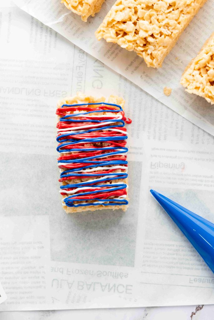 A rectangular Rice Krispies treat, perfect for 4th of July treats, is topped with red, white, and blue icing on parchment paper with printed text. A blue piping bag rests nearby, and more festive Rice Krispies treats are visible in the background.