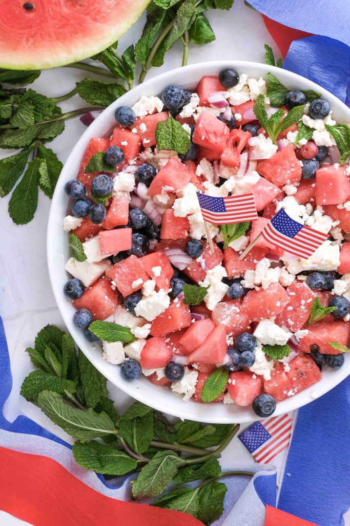 A bowl of Watermelon Blueberry Feta Salad with mint and tiny American flags sits amid red, white, and blue decorations. This Red White Blue Salad is perfect for a festive, patriotic Summer BBQ celebration.