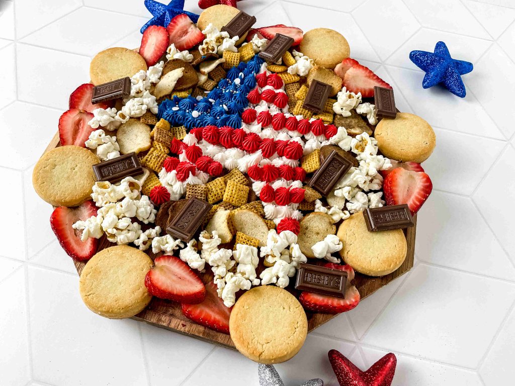 A festive charcuterie board displays cookies, strawberry slices, popcorn, and chocolate alongside a star-shaped centerpiece styled like the American flag—perfect for 4th of July snacks. Red and blue glitter stars add patriotic flair.