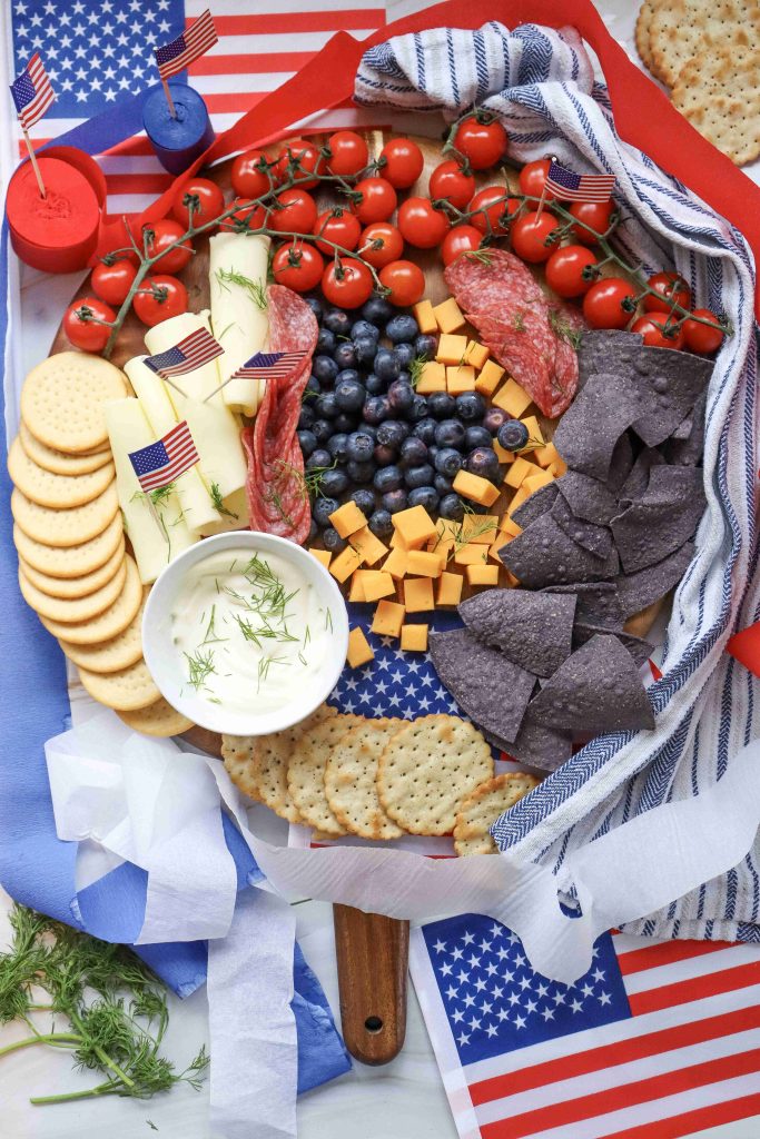 A 4th of July Charcuterie Board featuring crackers, cheese cubes, cherry tomatoes, blueberries, salami, blue chips, and dip. American flags decorate the food alongside stars-and-stripes napkins and patriotic ribbon in the background.