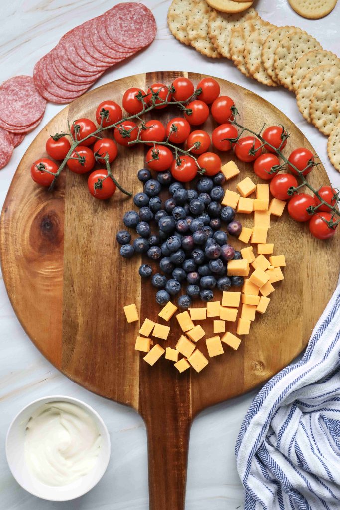 A 4th of July Charcuterie Board featuring a wooden board with grape tomatoes on the vine, blueberries, cheddar cubes, crackers, salami slices, and a bowl of creamy dip. A striped cloth sits beside this festive spread.
