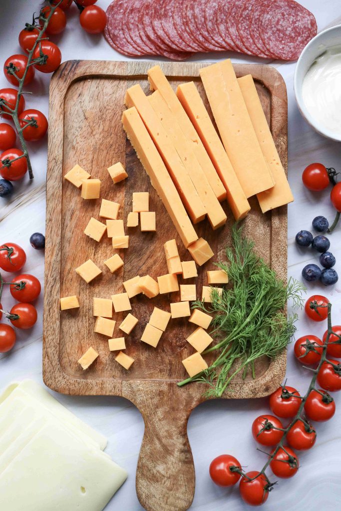 A festive 4th of July Charcuterie Board featuring a wooden board with sliced and cubed cheddar cheese, fresh dill, cherry tomatoes, blueberries, salami slices, a bowl of dip, and slices of white cheese.