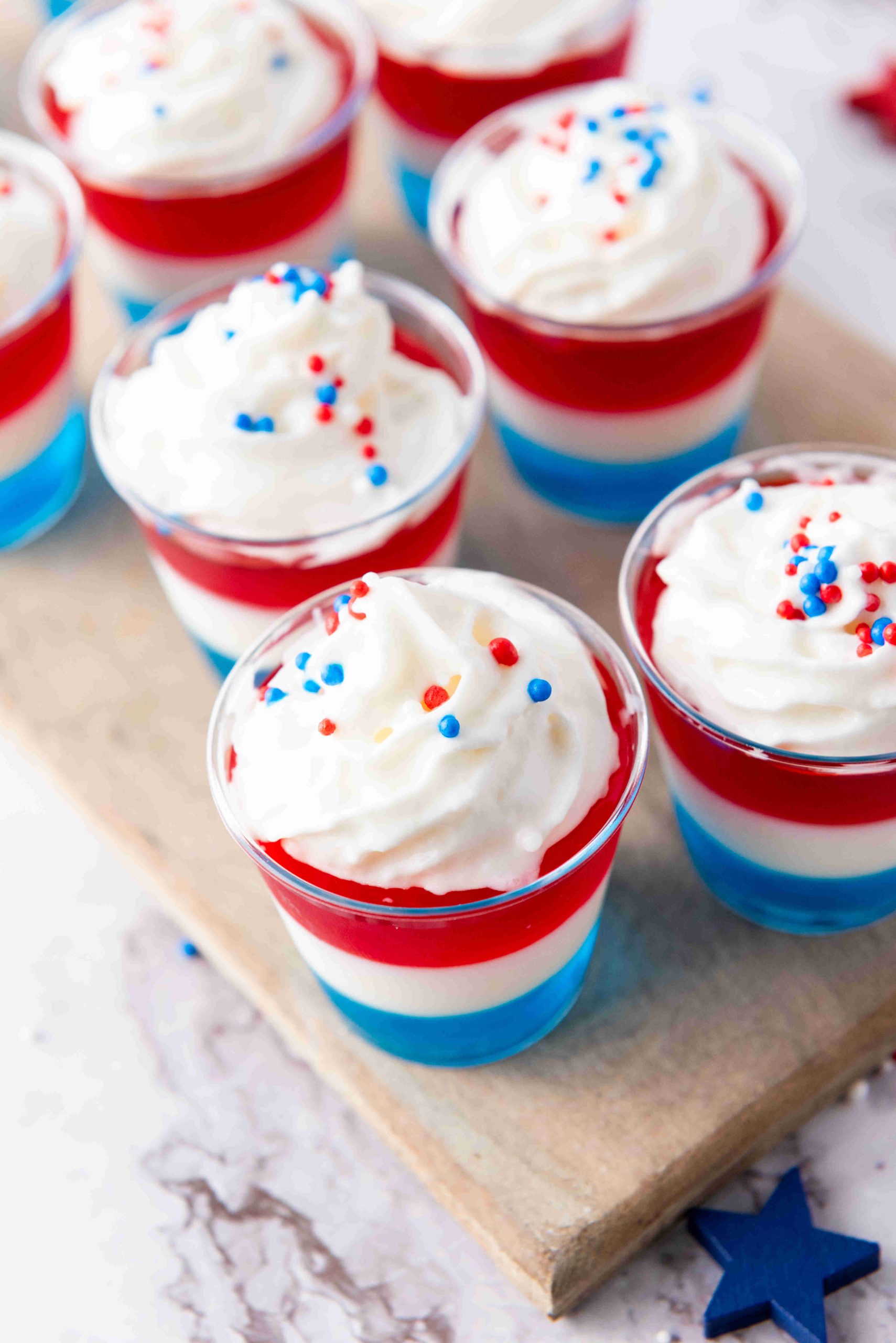 Small clear Jello Dessert Cups filled with layered red, white, and blue gelatin are topped with whipped cream and patriotic sprinkles. These festive treats make perfect Patriotic Desserts, artfully arranged on a wooden board.