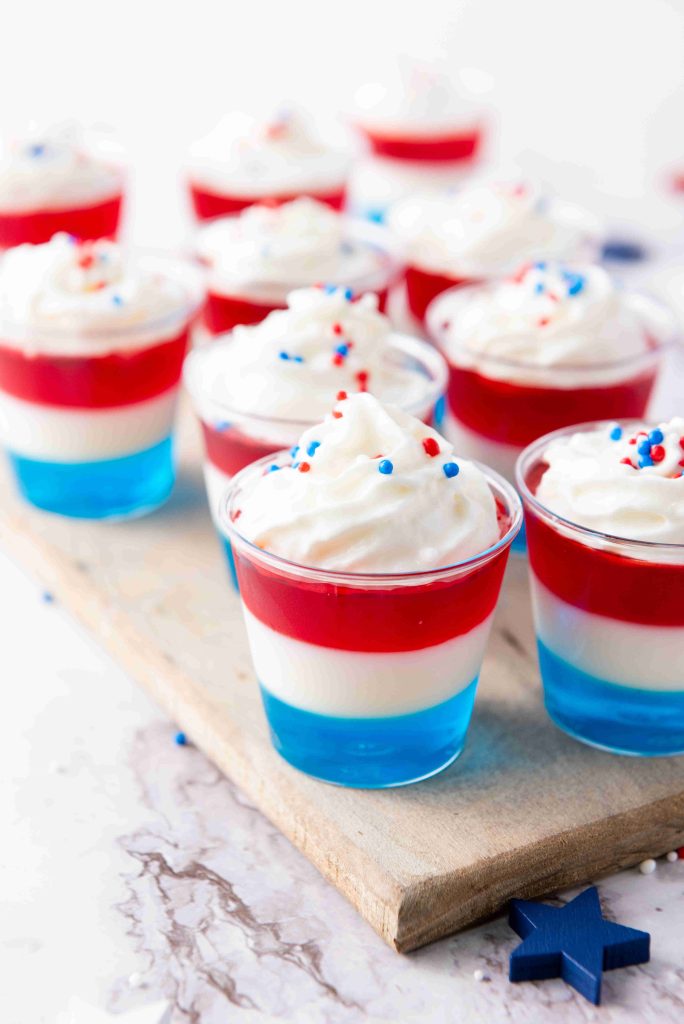 Several small cups of layered red, white, and blue 4th of July Jello Shots topped with whipped cream and festive sprinkles are arranged on a wooden board.