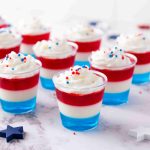 Small clear cups filled with layered red, white, and blue Jello Shots, topped with whipped cream and festive sprinkles. Decorative star-shaped confetti is scattered on the marble surface—perfect for your 4th of July celebration!.