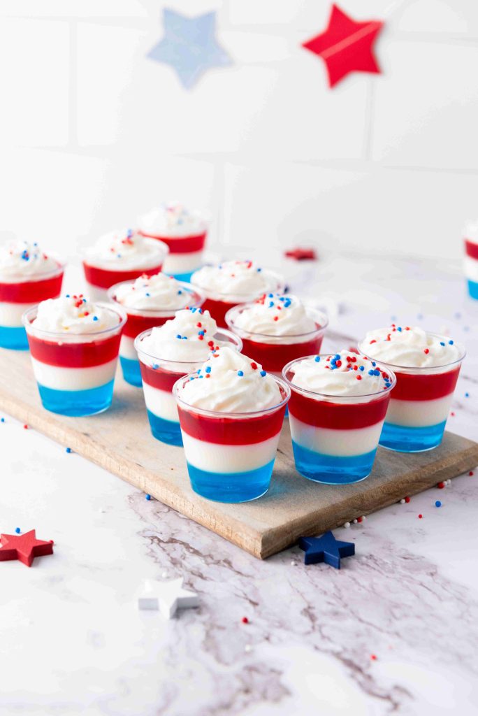 Small clear cups filled with layered red, white, and blue Jello Dessert Cups topped with whipped cream and festive sprinkles are arranged on a wooden board. Star decorations in the background complete these patriotic treats.