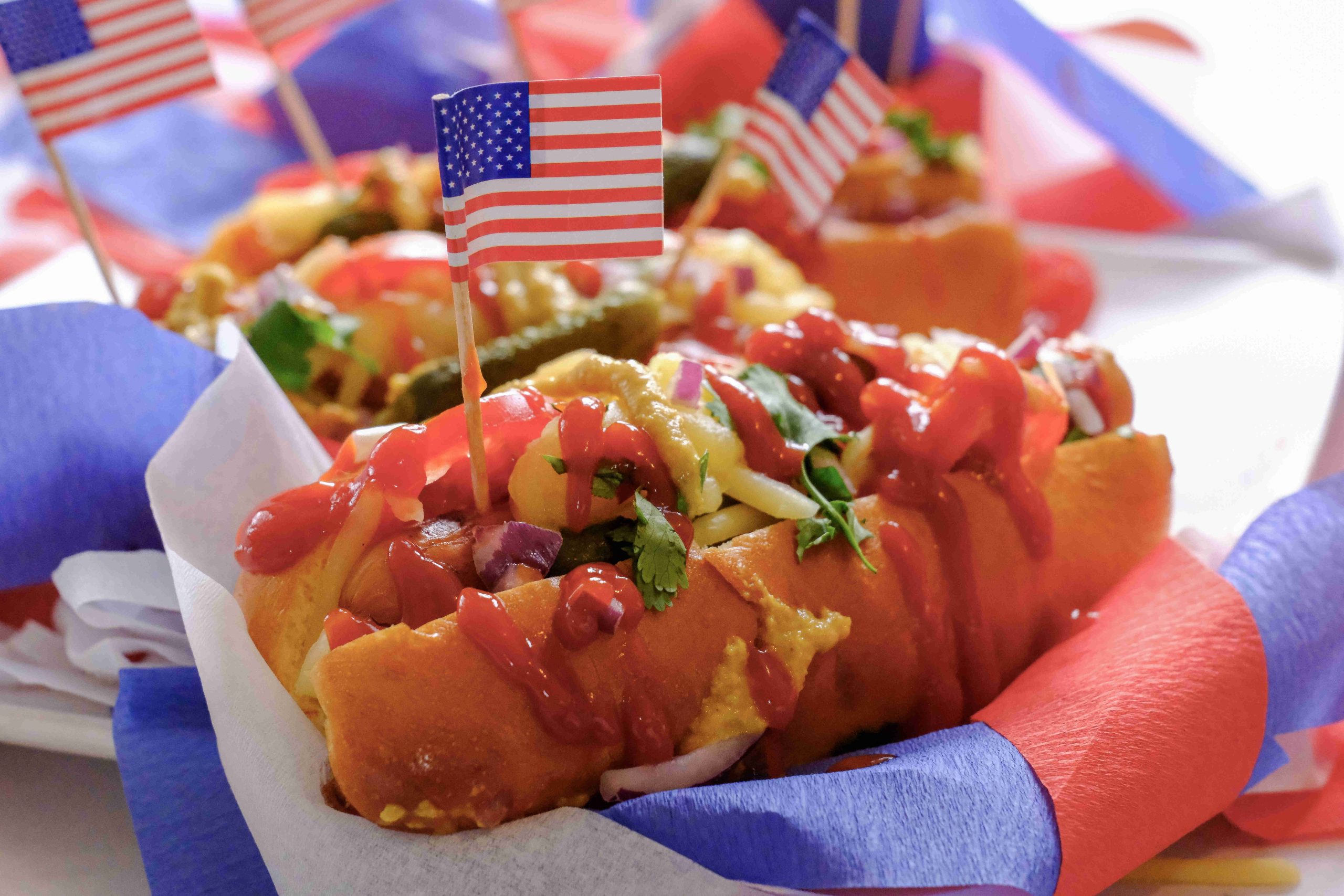 A close-up of American Hot Dogs topped with ketchup, mustard, onions, and herbs, each decorated with a small American flag toothpick, surrounded by red, white, and blue paper decorations.