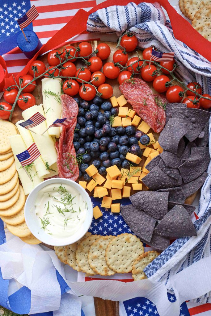A 4th of July Charcuterie Board with crackers, sliced cheese, cubed cheddar, blueberries, grape tomatoes, salami, blue corn chips, and dip, garnished with mini American flags and surrounded by festive napkins.
