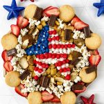 A round 4th of July Charcuterie Board showcases cookies, strawberries, popcorn, cereal, and chocolate pieces around the edge, with a star in the center decorated in red, white, and blue like the American flag. Sparkly stars add a festive touch.