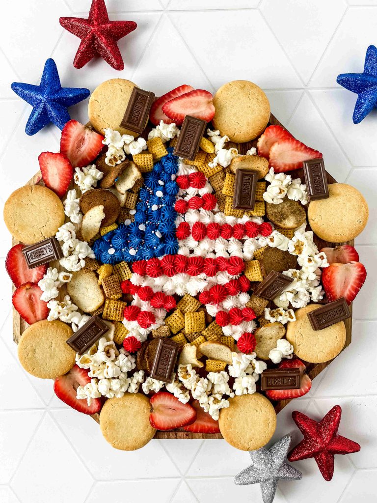 A round 4th of July Charcuterie Board showcases cookies, strawberries, popcorn, cereal, and chocolate pieces around the edge, with a star in the center decorated in red, white, and blue like the American flag. Sparkly stars add a festive touch.