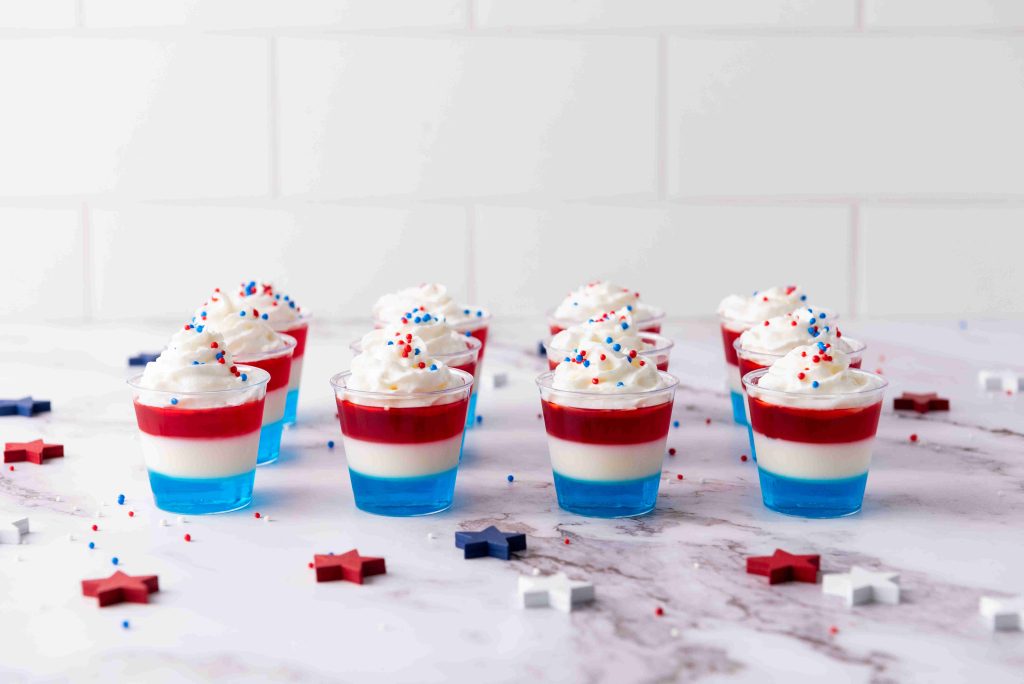 Nine small 4th of July Jello Shots with blue, white, and red layers are topped with whipped cream and festive sprinkles, arranged on a marble surface with star-shaped decorations nearby—perfect for your patriotic celebration.