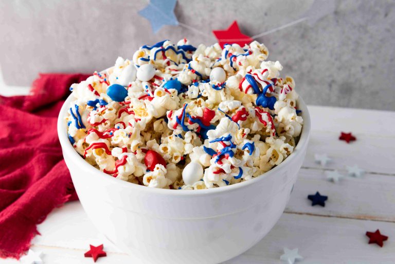 A white bowl filled with Patriotic Popcorn—topped with red, white, and blue M&Ms and drizzle—sits on a white wooden surface, surrounded by star-shaped confetti and a red cloth for a festive, patriotic touch.
