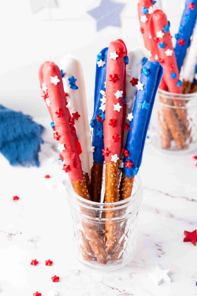 Patriotic Snacks shine with Pretzel Rods dipped in red, white, and blue candy coating, finished with star-shaped sprinkles and arranged in a glass jar. Red, white, and blue star sprinkles are scattered on a white surface.