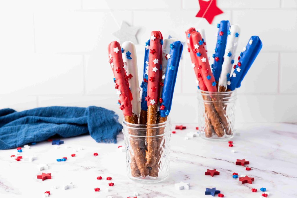 Glass jars hold Red White and Blue Pretzel Rods dipped in colorful coatings, decorated with star-shaped sprinkles. Scattered stars on the white marble surface complete this festive display of patriotic snacks.