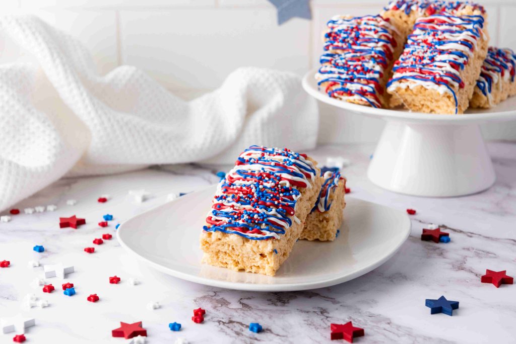 A plate with two festive 4th of July treats—Rice Krispie Treats decorated in red, white, and blue icing and sprinkles—sits on a marble counter. More decorated treats are on a cake stand in the background, surrounded by star-shaped confetti.