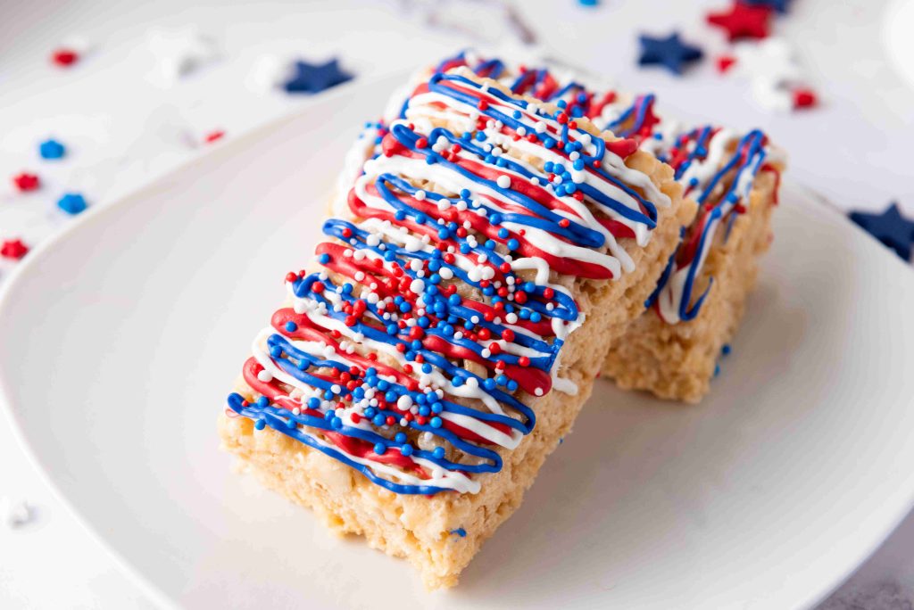 Two Rice Krispie Treats on a white plate, topped with red, white, and blue icing drizzle and sprinkles—perfect 4th of July desserts. Star-shaped confetti decorates the festive background.