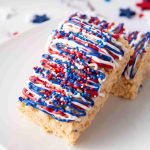 Two Rice Krispie Treats on a white plate, topped with red, white, and blue icing drizzle and sprinkles—perfect 4th of July desserts. Star-shaped confetti decorates the festive background.