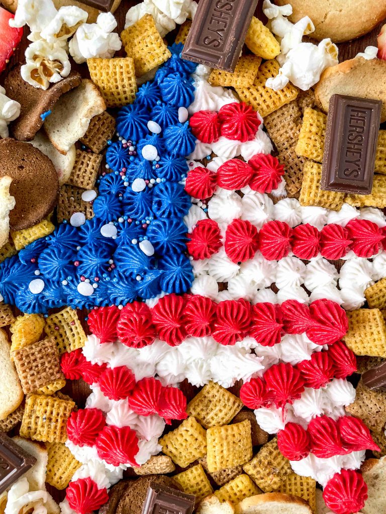 A variety of snacks surround a large star-shaped cake decorated with red, white, and blue frosting and sprinkles, creating the perfect 4th of July charcuterie board with cereal, cookies, popcorn, and Hershey’s chocolate bars.