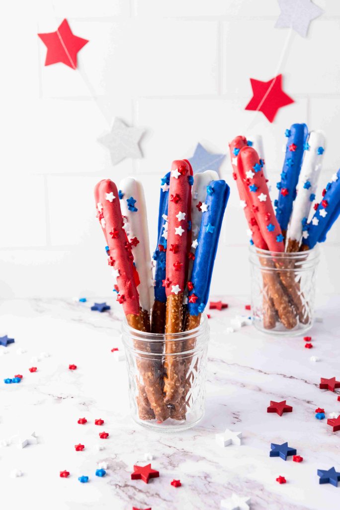 A glass jar displays Red White and Blue Pretzel Rods dipped in candy coating and topped with star sprinkles—festive Patriotic Snacks perfect for celebrations. Another jar and star confetti decorate the white surface in the background.
