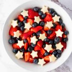 A white bowl filled with sliced strawberries, whole blueberries, and small star-shaped pieces of white fruit creates a festive red white and blue fruit salad, beautifully arranged on a marble surface.