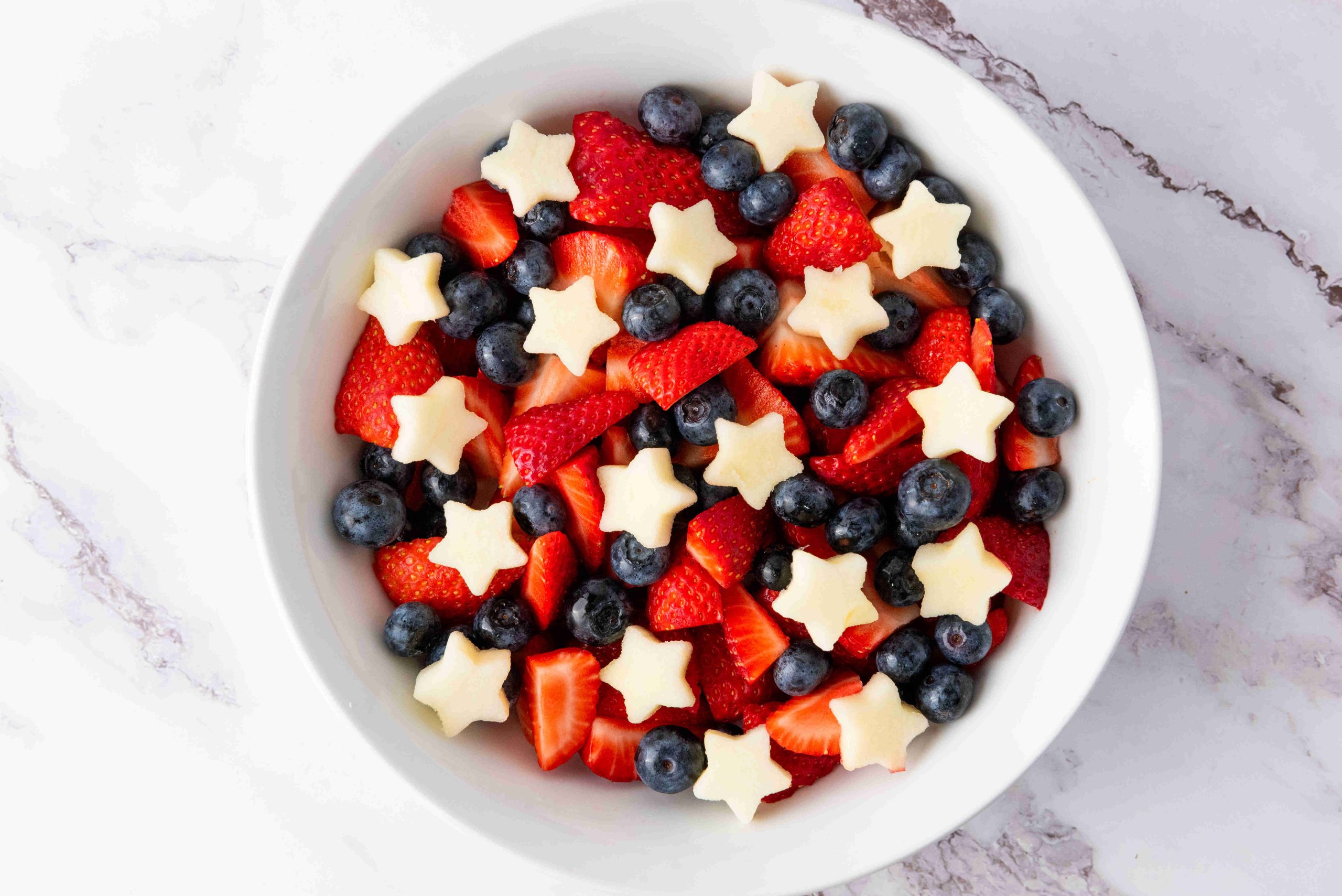 A white bowl filled with sliced strawberries, whole blueberries, and small star-shaped pieces of white fruit creates a festive red white and blue fruit salad, beautifully arranged on a marble surface.