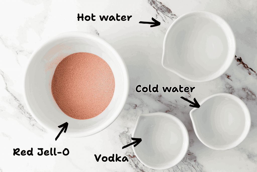 A bowl of red Jell-O powder and three bowls labeled hot water, cold water, and vodka are arranged on a marble surface—perfect for making 4th of July Vodka Jello Shots. Each bowl is clearly marked with an arrow.