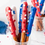 Red White and Blue Pretzel Rods dipped in chocolate and topped with star-shaped sprinkles stand upright in clear glass jars, creating a festive, patriotic display.