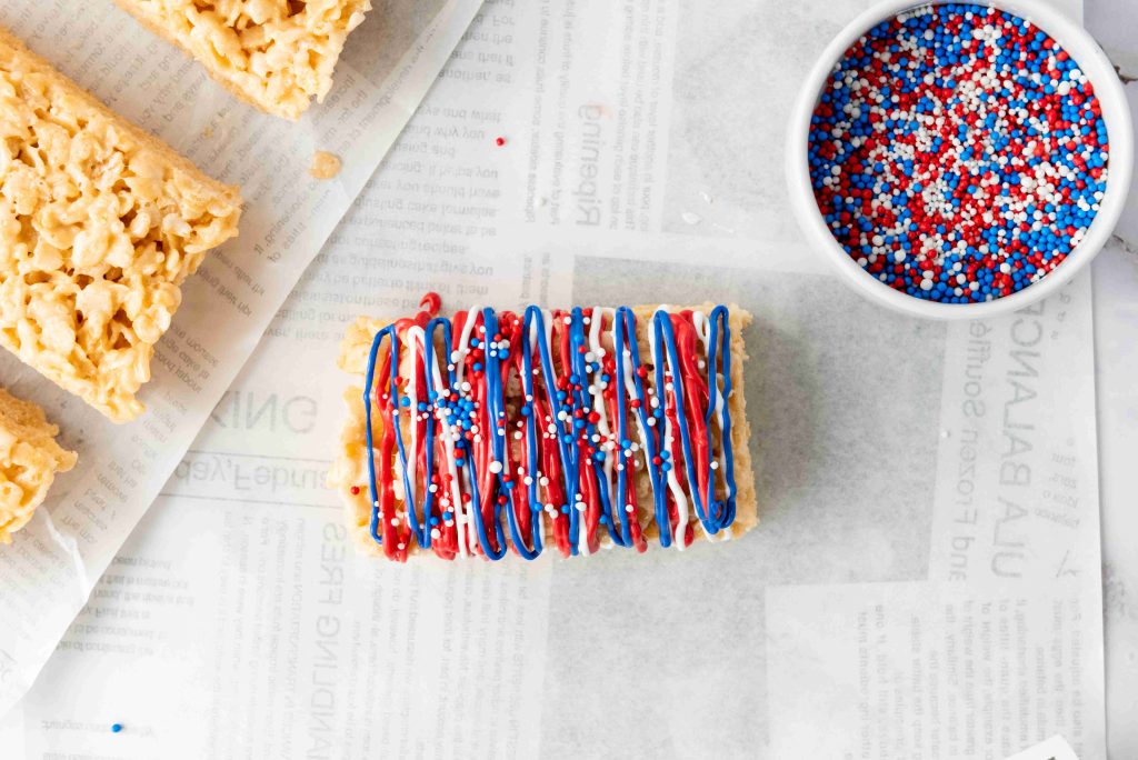 A Rice Krispie Treats dessert is topped with red, white, and blue icing and sprinkles for a festive 4th of July look, placed on parchment paper next to a bowl of matching sprinkles and plain treats.