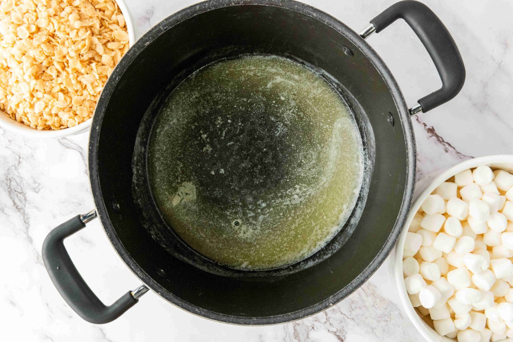 A large black pot with melted butter sits on a marble countertop, surrounded by bowls of cornflakes and mini marshmallows, perfect for whipping up festive 4th of July desserts or creative Rice Krispie Treats.
