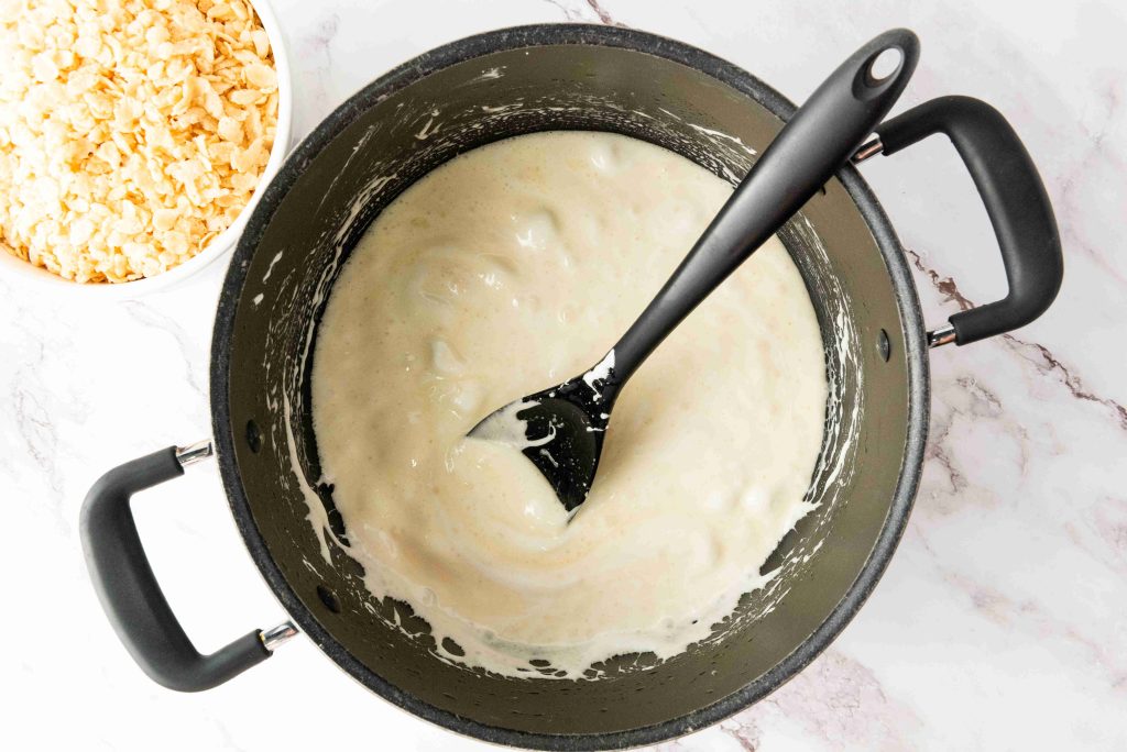 A black pot filled with melted marshmallows is being stirred with a black spatula, ready for delicious Rice Krispie Treats. A bowl of rice cereal sits nearby on a white marble surface—perfect for festive 4th of July treats.