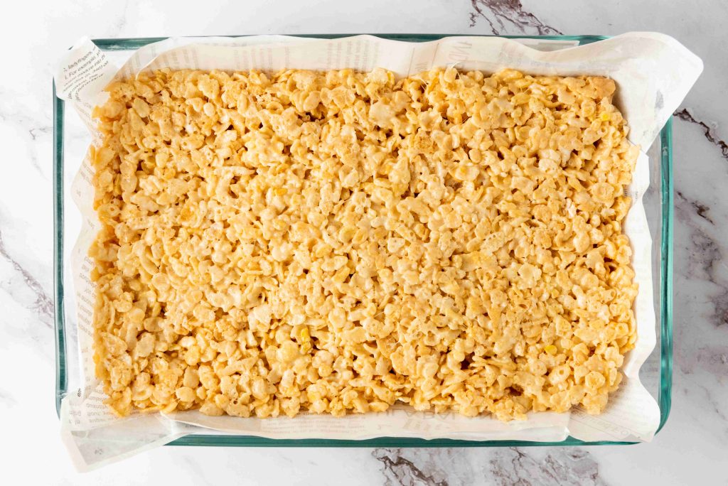 A rectangular glass baking dish lined with parchment paper, filled with a pressed layer of golden Rice Krispie Treats—a perfect base for festive 4th of July desserts—sits on a white marble surface.