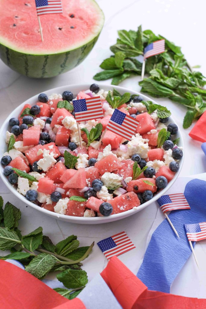 A bowl of Watermelon Blueberry Feta Salad, decorated with small American flags, sits on a table with fresh mint, a watermelon, and red and blue napkins nearby—the perfect Red White Blue Salad for your next Summer BBQ.