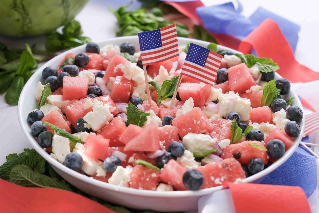 A festive Red White Blue Salad of watermelon cubes, blueberries, feta cheese, and mint leaves in a white bowl, decorated with two small American flags and surrounded by red, white, and blue ribbons—perfect for your Summer BBQ.