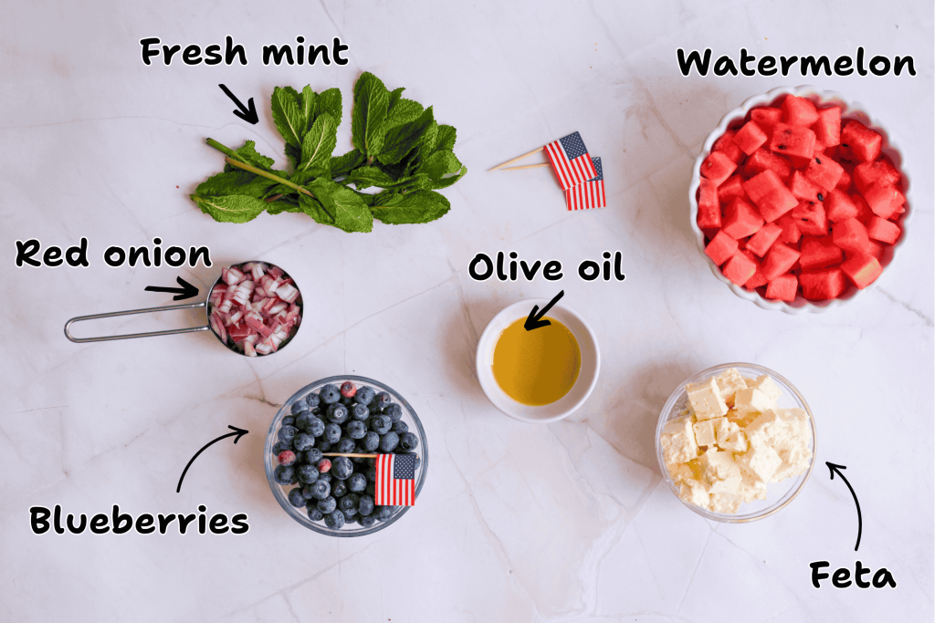 Ingredients for a Summer BBQ Red White Blue Salad are arranged on a white surface: fresh mint, diced red onion, blueberries, cubed watermelon, olive oil, and feta cheese—each labeled with handwritten text and some with small American flag picks.
