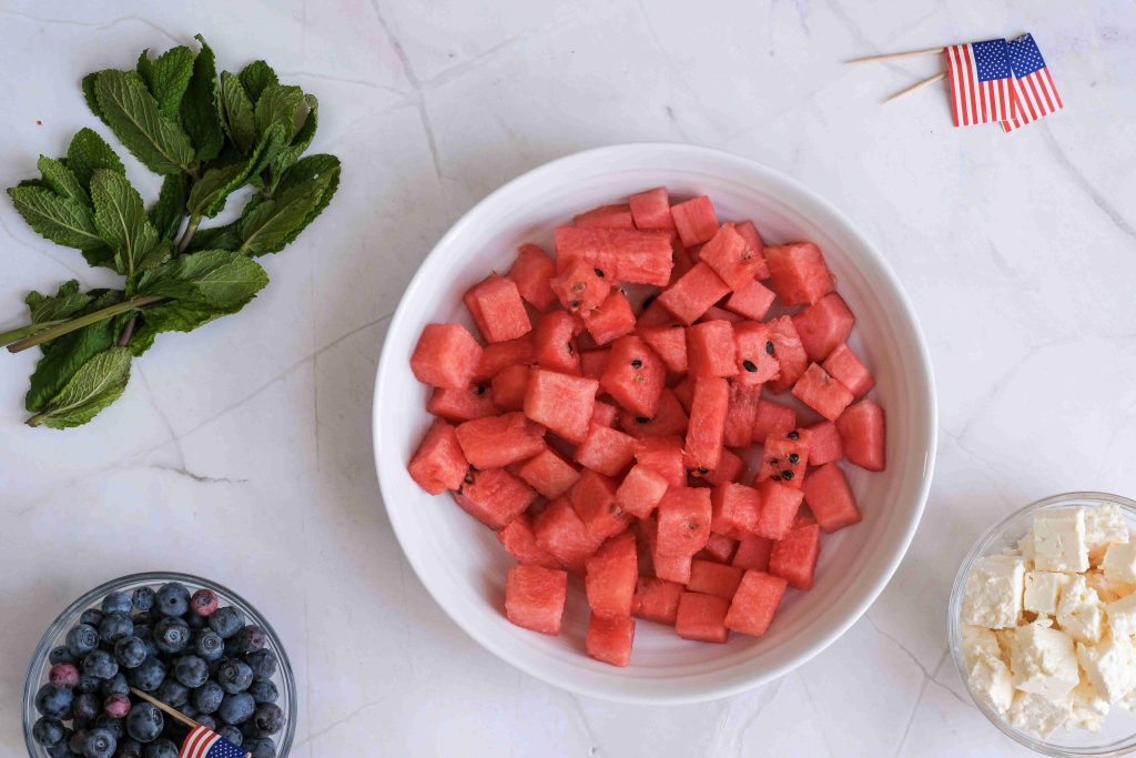 A white bowl filled with Watermelon Blueberry Feta Salad sits on a marble surface, surrounded by fresh mint, a bowl of blueberries, feta cheese, and small American flag toothpicks—perfect for your Summer BBQ or Red White Blue Salad spread.