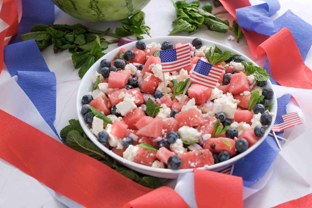 A bowl of Watermelon Blueberry Feta Salad is decorated with small American flags and surrounded by red, white, and blue ribbons, making it the perfect festive treat for your Summer BBQ.