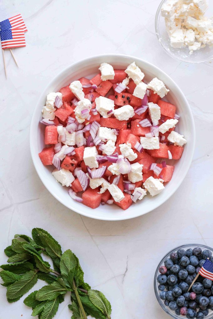 A refreshing Red White Blue Salad features watermelon cubes, crumbled feta, chopped red onions, mint leaves, blueberries, and a small American flag—perfect for your next Summer BBQ.