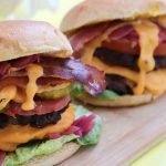 Two American Burgers on a wooden board, topped with melted cheese, bacon, pickles, lettuce, tomato, onions, and sauce, with a bowl of pickled onions in the background.