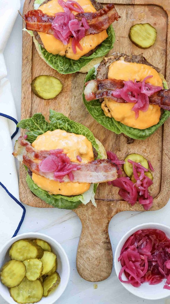 Three bacon cheeseburgers topped with pickled onions sit on lettuce leaves on a wooden board, surrounded by sliced pickles and extra pickled onions—an irresistible American burger recipe.