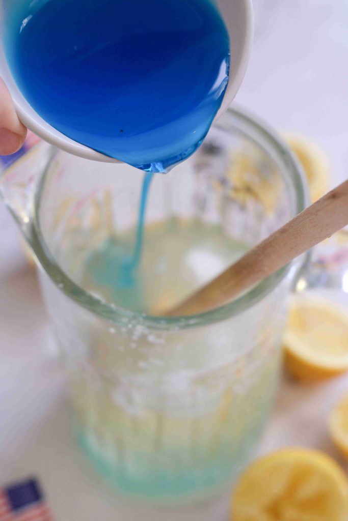 A hand pours bright blue liquid from a small white cup into a glass pitcher filled with a pale yellow Blue Lemonade Mocktail, with a wooden spoon inside and halved lemons nearby—perfect as a kid-friendly drink.