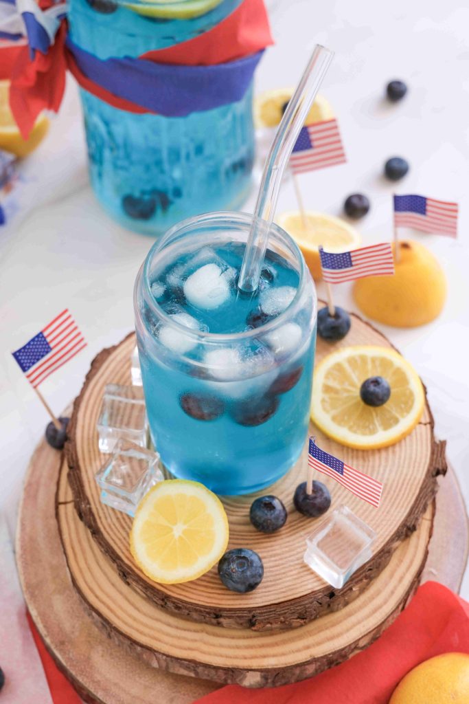 A glass of sparkling blue lemonade mocktail with ice cubes, blueberries, and a clear straw is garnished with lemon slices. Small American flags, blueberries, and lemons decorate the wooden stand for a festive Fourth of July drink scene.
