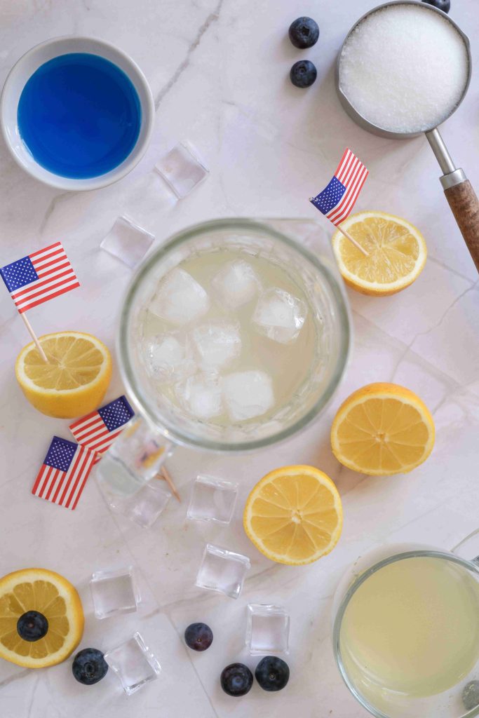 A top view of a festive Blue Lemonade mocktail with ice, surrounded by lemon slices, blueberries, small American flags, a bowl of blue liquid, sugar, and scattered ice cubes—perfect as a Fourth of July drink on a marble surface.